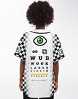 Black-and-white checkerboard EDM baseball jersey on a woman standing with her back facing forward. The back shows a large green cyclops eye, infinity symbol, geometric shapes, and a full eye-chart parody with words like WUB, WOOK, LASER, RIDDIM, and CYCLOPS. Sleeves have matching green-eye patches.